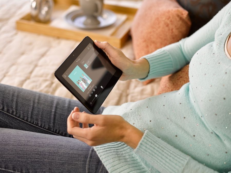 A person sitting on a bed holds a smart home control tablet displaying a music interface.