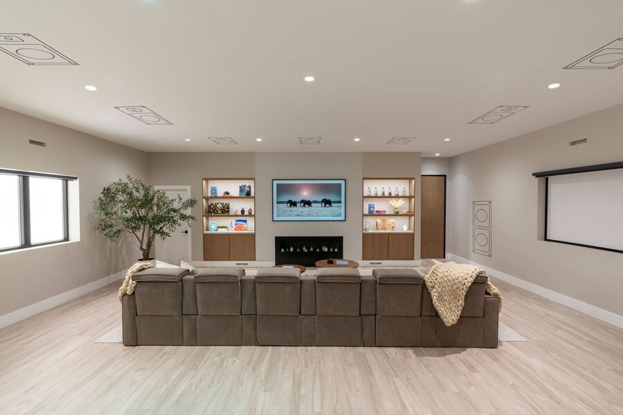 Modern home theater with a large sectional sofa facing a wall-mounted TV, built-in shelving with accent lighting, and hidden speakers integrated into the ceiling and walls.