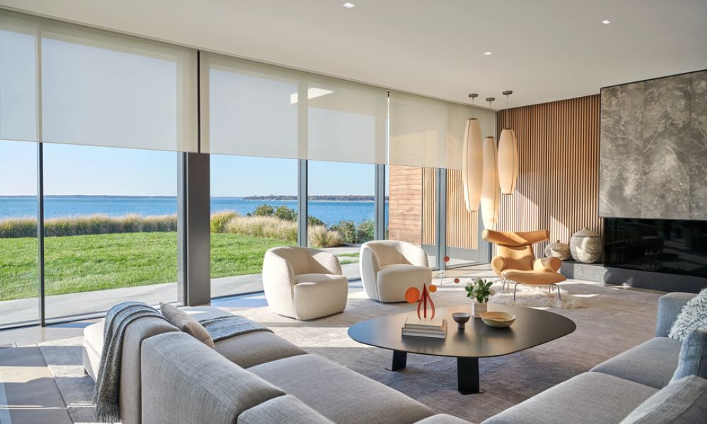 A modern living room with floor-to-ceiling glass windows, neutral furnishings, a view of a grassy yard and water, wood paneling, and contemporary decor.