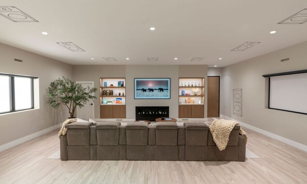 Modern home theater with a large sectional sofa facing a wall-mounted TV, built-in shelving with accent lighting, and hidden speakers integrated into the ceiling and walls.