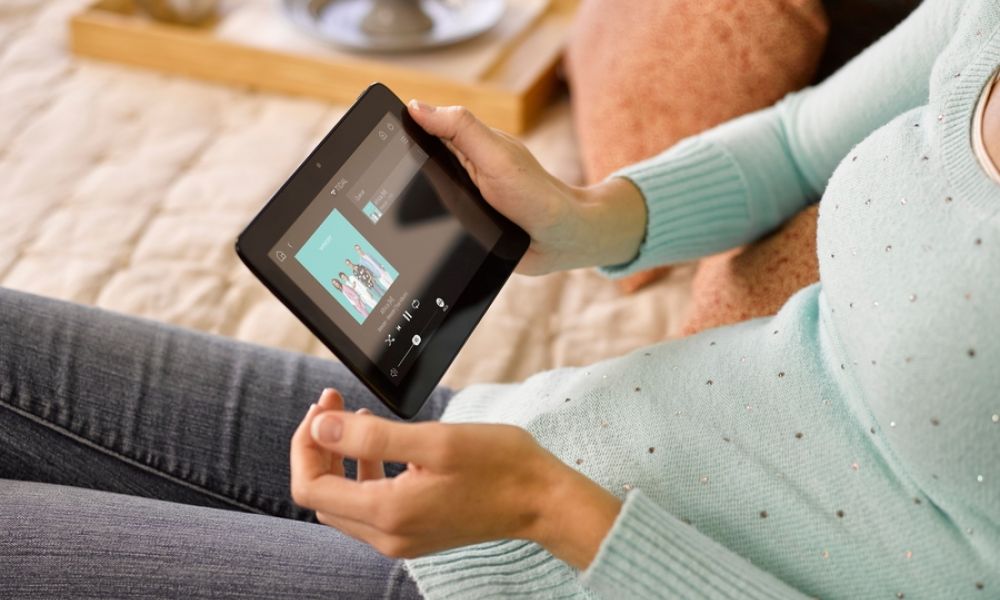 A person sitting on a bed holds a smart home control tablet displaying a music interface.