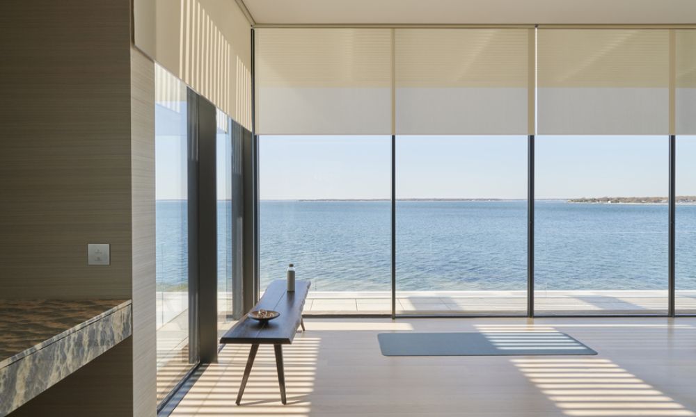 Motorized Lutron shades partially lowered over floor-to-ceiling windows in a modern waterfront home with ocean views.  Social Media Bl
